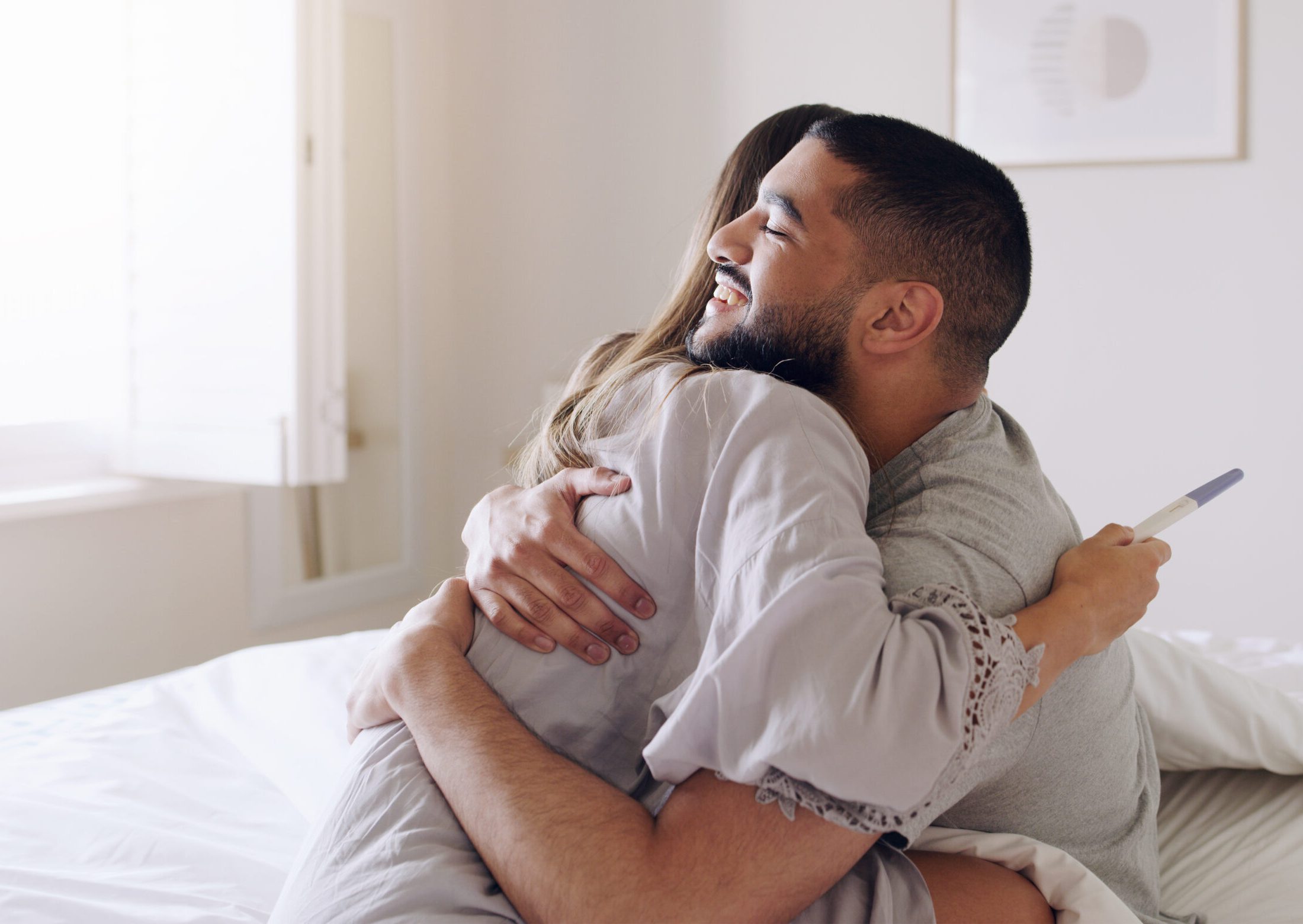 Couple, hug and happy for pregnancy test in bedroom, home and support to celebrate good news. Excited man, woman and partner hugging in celebration of pregnant results, future family or ivf fertility Couple, hug and happy for pregnancy test in bedroom, home and support to celebrate good news. Excited man, woman and partner hugging in celebration of pregnant results, future family or ivf fertility.